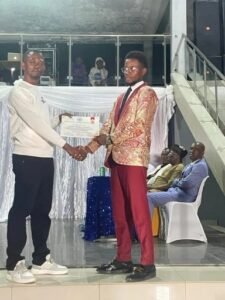 Award ceremony: a man in a light sweater presents a certificate to a recipient in a gold-patterned jacket and red pants on stage, with seated guests in the background.