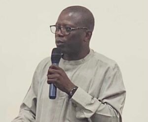 | The Calabash Newspaper Man wearing a light traditional shirt and glasses, speaking into a handheld microphone.