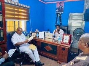 Three men in a blue-walled office sit around a wooden desk; one reads a book, another consults a laptop, while the third speaks and gestures.