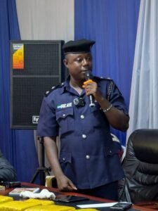 Nigerian police officer in a dark blue uniform speaks into a handheld microphone at an indoor event, with a speaker behind him and blue curtains in the background.