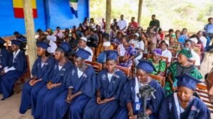 Graduates in blue gowns sit in front under a shelter while a crowd watches the ceremony.