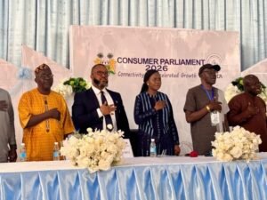 Six diverse delegates stand with hands over their hearts behind a table with white floral arrangements at a conference stage reading 'Consumer Parliament 2026' in the background.
