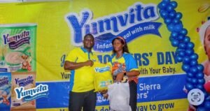 Two people pose with Yumvita infant cereal samples at a promotional event in front of a bright yellow Yumvita banner.