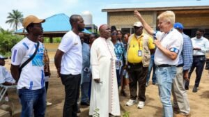 Group of people outdoors listening to a speaker with a microphone, a priest in a white robe at center, as others stand nearby near a brick building and palm tree.