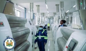 Industrial textile plant with rows of large white machines and overhead ducts; workers in blue uniforms and safety gear walk the aisle.