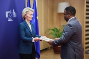Woman in a teal blazer hands a document to a man in a gray suit, EU flags in the background.