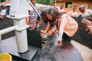 Woman in an orange safety vest lifts the hatch on a large black water tank as onlookers watch at a community water site.