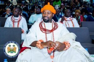 | The Calabash Newspaper Man in a white ceremonial robe with an orange turban and multiple red bead necklaces sits on stage at a formal event, audience behind him, seal of the Office of the President visible.