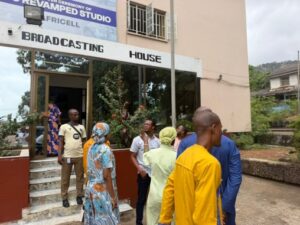People in colorful clothing stand outside the Broad Casting House entrance of a broadcast studio building.