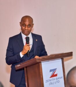 Man in a pinstripe suit speaks into a handheld microphone at a wooden podium, with a Zenith Bank logo on the lectern.