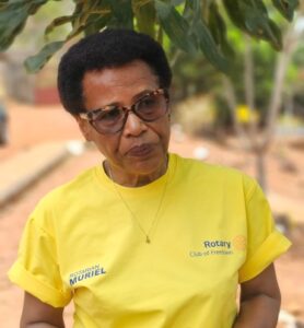 Portrait of an older woman wearing a yellow Rotary Club shirt with the name Muriel, sunglasses, outdoors under trees.