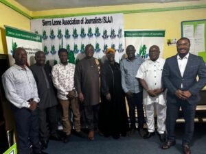 Group of eight adults posing for a photo in a room with Sierra Leone Association of Journalists banners in the background.