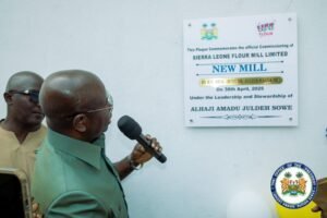 Man in a green suit speaks into a microphone beside a plaque announcing a new Sierra Leone Flour Mill (NEW MILL).