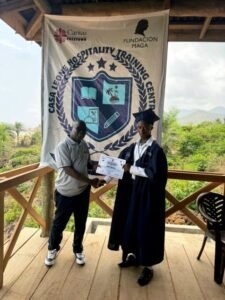 | The Calabash Newspaper Graduate in a cap and gown receives a certificate from a man on a wooden deck, with a Hospitality Training Center banner behind them.