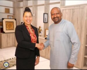 Two professionals in a modern office shake hands and smile at the camera. The woman wears a black blazer and a red floral blouse; the man wears a light gray traditional outfit.