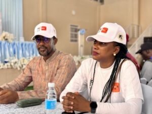 | The Calabash Newspaper Two attendees wearing white caps sit at a table during an indoor event; man in a patterned shirt and woman with braids in a white top, listening attentively.