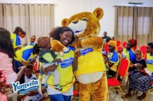 Woman holding a baby hugs a bear mascot in a yellow Yumvita shirt at a children’s event with attendees seated in red chairs in a hall.