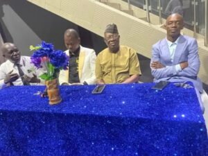 Four men sit at a long table covered with a blue glittery cloth, with a potted purple flower centerpiece in the middle.
