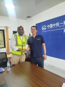 Two men shake hands in an office: one in a neon safety vest, the other in a navy polo, standing in front of a blue wall with a China Railway First Group logo.
