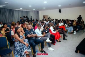 Audience seated in a conference hall listening to a presentation; diverse attendees in formal and casual attire.