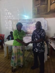 Man in a floral shirt presents a certificate to a woman in a green shirt at an indoor ceremony with a banner in the background.
