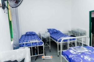 Three single hospital beds with blue patterned bedding and small bedside carts in a bright white ward, with a window and curtain and a wall fan above.