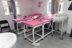 Treatment room with three pink padded examination chairs on wheeled metal frames by tiled walls and windows.