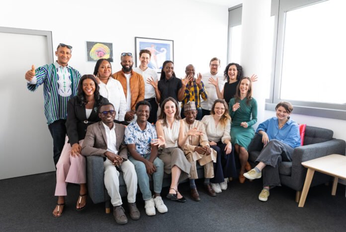Wiener Zeitung Engages African Journalists Group of diverse colleagues posing together in a bright office, smiling and waving at the camera.