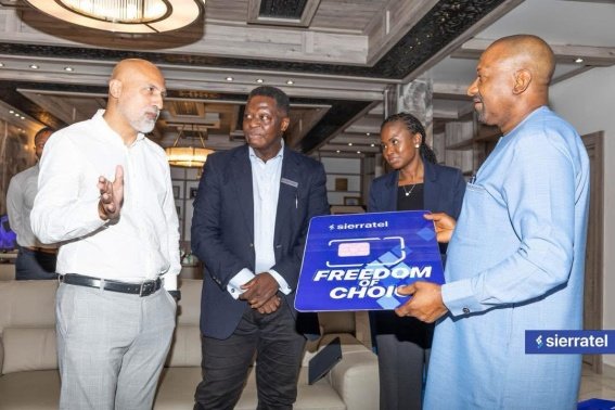 Four professionals in a modern lounge pose for a photo; one man in blue traditional attire holds a blue sign reading 'Freedom of Choice' with the Sierratel logo, while two others in formal wear listen.