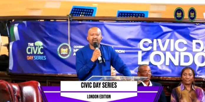 Man in a blue shirt speaks into a microphone at a podium during a Civic Day Series London edition event, with a blue banner in the background.