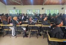Over 450 Returned Migrants Graduate from IOM Supported TVET Programme Group of graduates in black gowns and mortarboards seated at desks inside a bright classroom or lecture hall, some taking photos, with large windows along the wall and ceiling fans above.