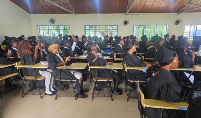 Group of graduates in black gowns and mortarboards seated at desks inside a bright classroom or lecture hall, some taking photos, with large windows along the wall and ceiling fans above.