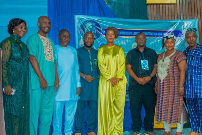 Nine people in colorful traditional outfits posing for a group photo at an event, with a blue banner behind them.