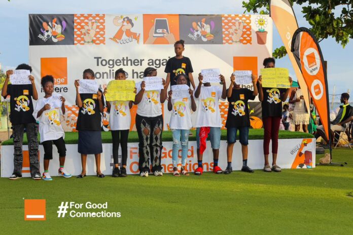 Group of diverse children standing on a stage outdoors, each holding handwritten signs during a community event with orange-themed banners in the background. The banner behind them reads 'Together again online' and there is a '#For Good Connections' graphic at the front left.