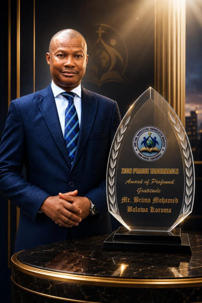 Man in a navy suit stands beside a tall glass award trophy on a marble table, the plaque inscribed with Zion Praise Tabernacle and 'Award of Profound Gratitude' for Mr. Brina Mohamed Baliwa Koroma.