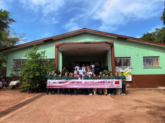 Group of people standing in front of a green building on a sunny day, holding a banner for a community event.