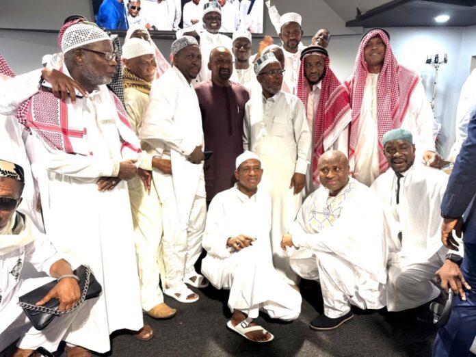 Group of Muslim men in white traditional attire posing for an indoor group photo, with several kneeling in front and others standing behind.