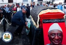 President Bio, First Lady Join Nation in Honouring Late APC Chairman Alhaji Minkailu Mansaray State funeral scene with a coffin draped in ornate cloth beside a large portrait of a man, as a man lays a bouquet amid a crowd of onlookers and photographers.