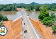 Manowa Bridge Opens New Era of Connectivity and Trade Across Kailahun District Aerial view of a straight two-lane bridge crossing a wide muddy river in a rural area, with people walking along the road and red dirt banks on both sides; government seal in the lower-left corner.