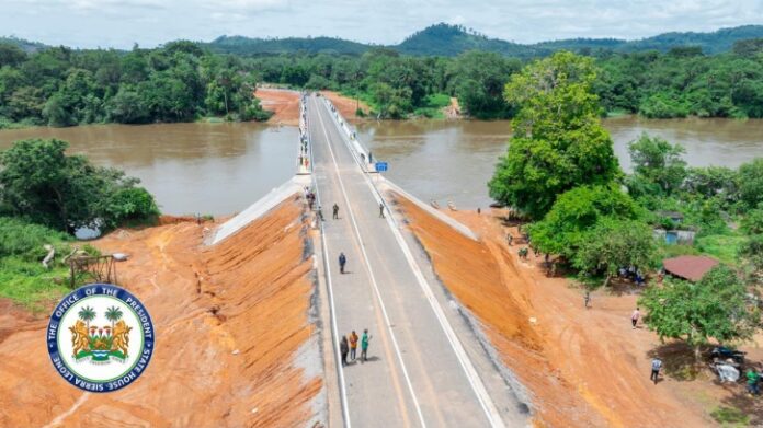 Aerial view of a straight two-lane bridge crossing a wide muddy river in a rural area, with people walking along the road and red dirt banks on both sides; government seal in the lower-left corner.