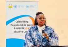 Gender Minister Champions Women’s Role in Peacebuilding, Calls for Greater Investment in Sustaining Peace Woman in a blue patterned outfit speaks into a handheld microphone at a conference, with banners about Peacebuilding Week and UN PBF in Sierra Leone in the background.