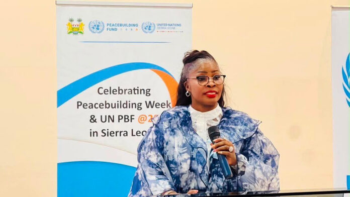 Woman in a blue patterned outfit speaks into a handheld microphone at a conference, with banners about Peacebuilding Week and UN PBF in Sierra Leone in the background.