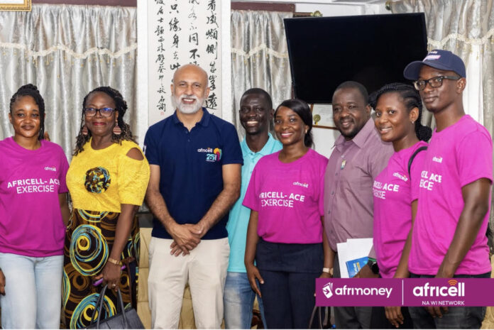 Group of eight people posing for a photo at a community event; several wear pink 'Africell-Ace Exercise' shirts, with Afrimoney and Africell logos visible on a banner/bracelet.