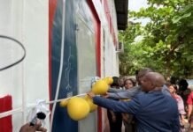 FirstBank Hands Over Renovated Solar-Powered Science and Computer Labs to Albert Academy Group of people in a ceremonial unveiling, pulling a rope with yellow balloons attached beside a blue-red wall.