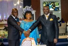 Zion Praise Tabernacle Celebrates 14 Years of Faith, Growth and Impact Three formally dressed adults on a stage shake hands over a table with a cake featuring a cross centerpiece.