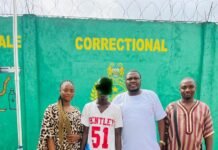 Rise Charity Foundation Secures Release of Liberian National from Bo Correctional Centre Group of four adults standing in front of a green wall reading 'Correctional' with barbed wire; a woman in leopard-print dress with a patterned bag, a boy in a white 'Bentley 51' shirt, a man in white shirt and red pants, and a man in a striped robe with a crossbody bag.