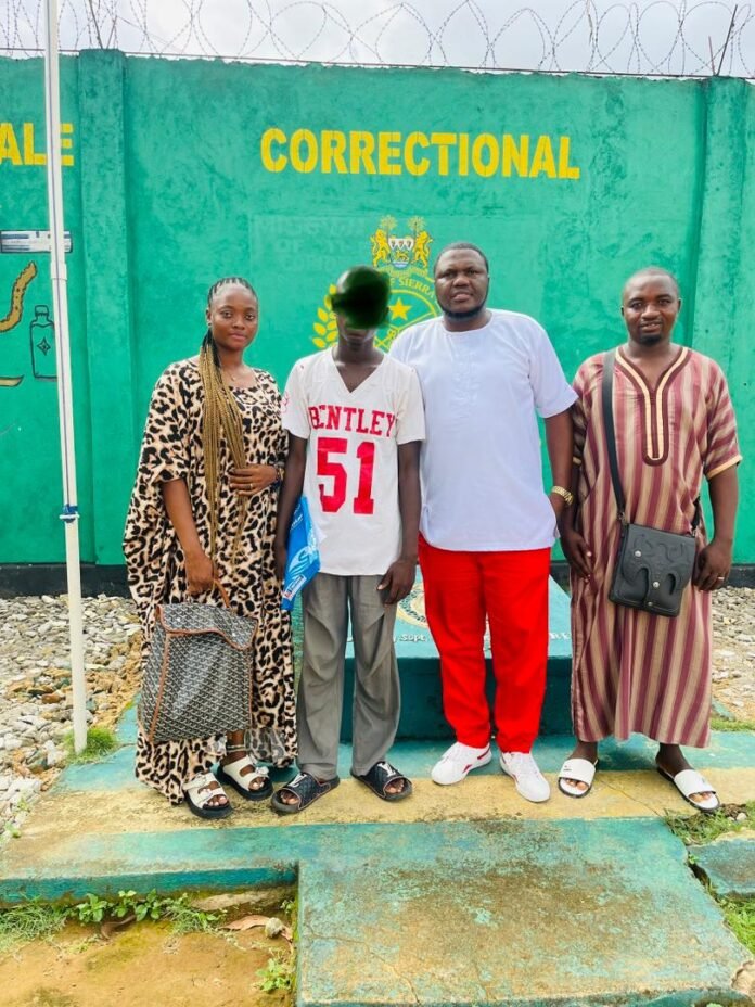 Group of four adults standing in front of a green wall reading 'Correctional' with barbed wire; a woman in leopard-print dress with a patterned bag, a boy in a white 'Bentley 51' shirt, a man in white shirt and red pants, and a man in a striped robe with a crossbody bag.