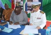 EU Strengthens Sierra Leone’s Naval Capacity With RHIBs and Forensic Equipment Two men sign documents at a table during a formal ceremony, one in a gray traditional outfit and the other in a white naval uniform, with onlookers in the background.
