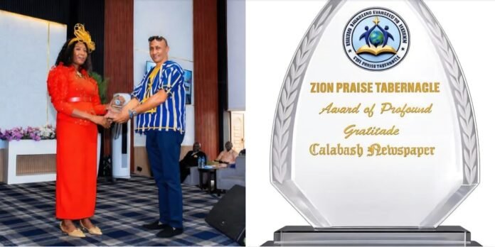 Two people on a stage exchange a trophy during an award ceremony; woman in red dress and man in blue striped shirt pose for the photo.