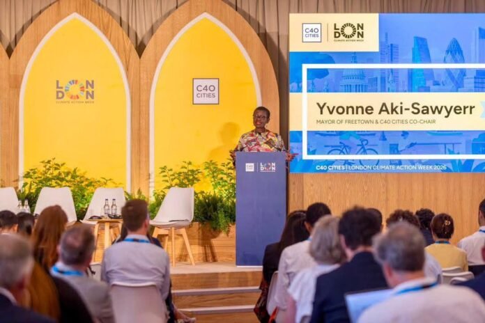 Speaker at a podium during London Climate Action Week, with C40 Cities branding and a large screen showing Yvonne Aki-Sawyerr.